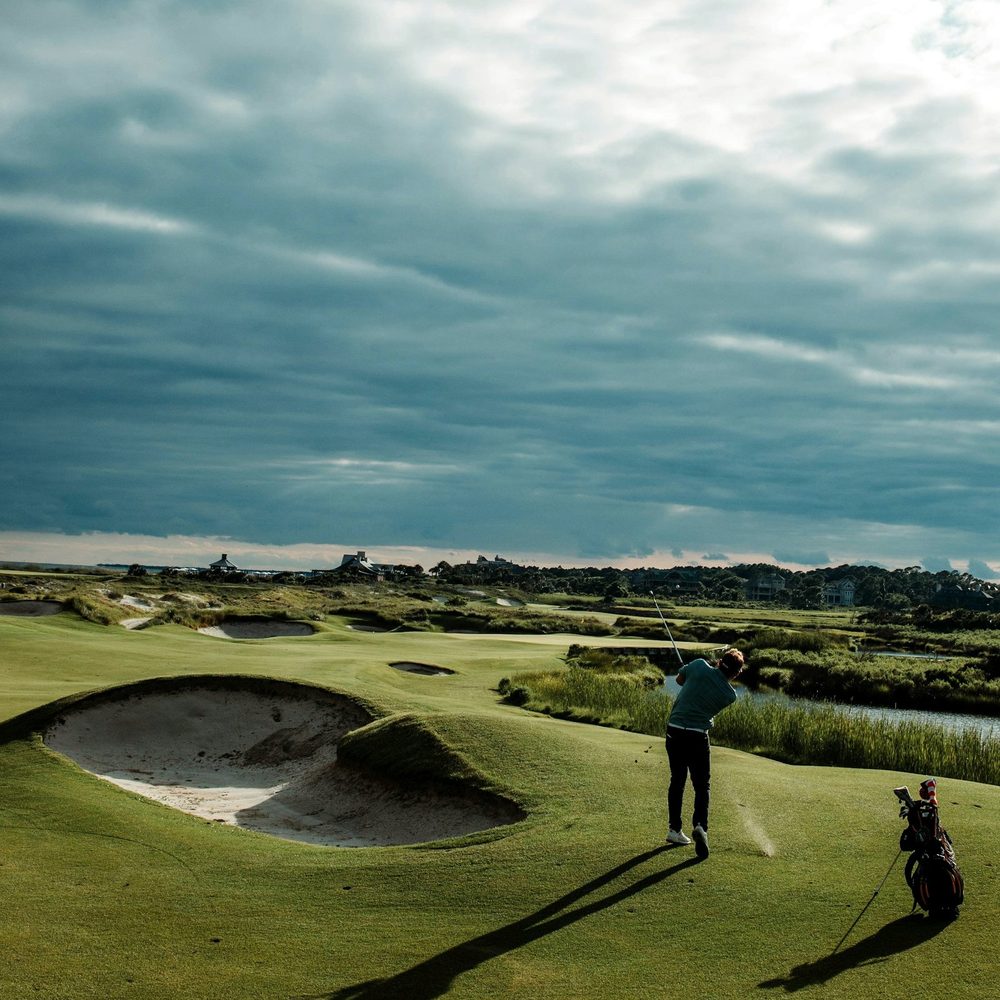 A golfer playing from beside a bunker on a links course under a wide sky